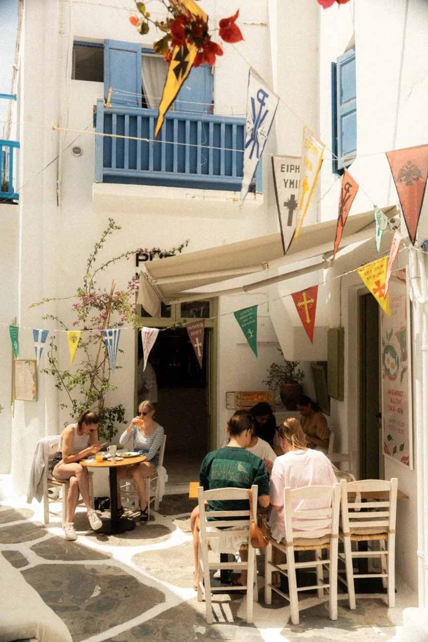 Outdoor terrace of Mykonos Brunch Club on a cobbled Old Town alley — colourful pennants, whitewashed walls, all-day breakfast Mykonos