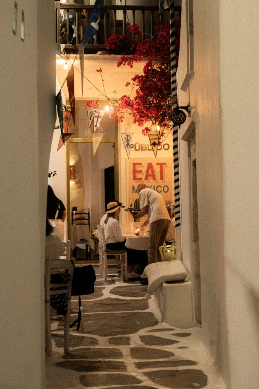Entrance to Pūblico restaurant viewed through a narrow Mykonos alley — EAT MEXICO sign glowing, bougainvillea overhead, waiter at work